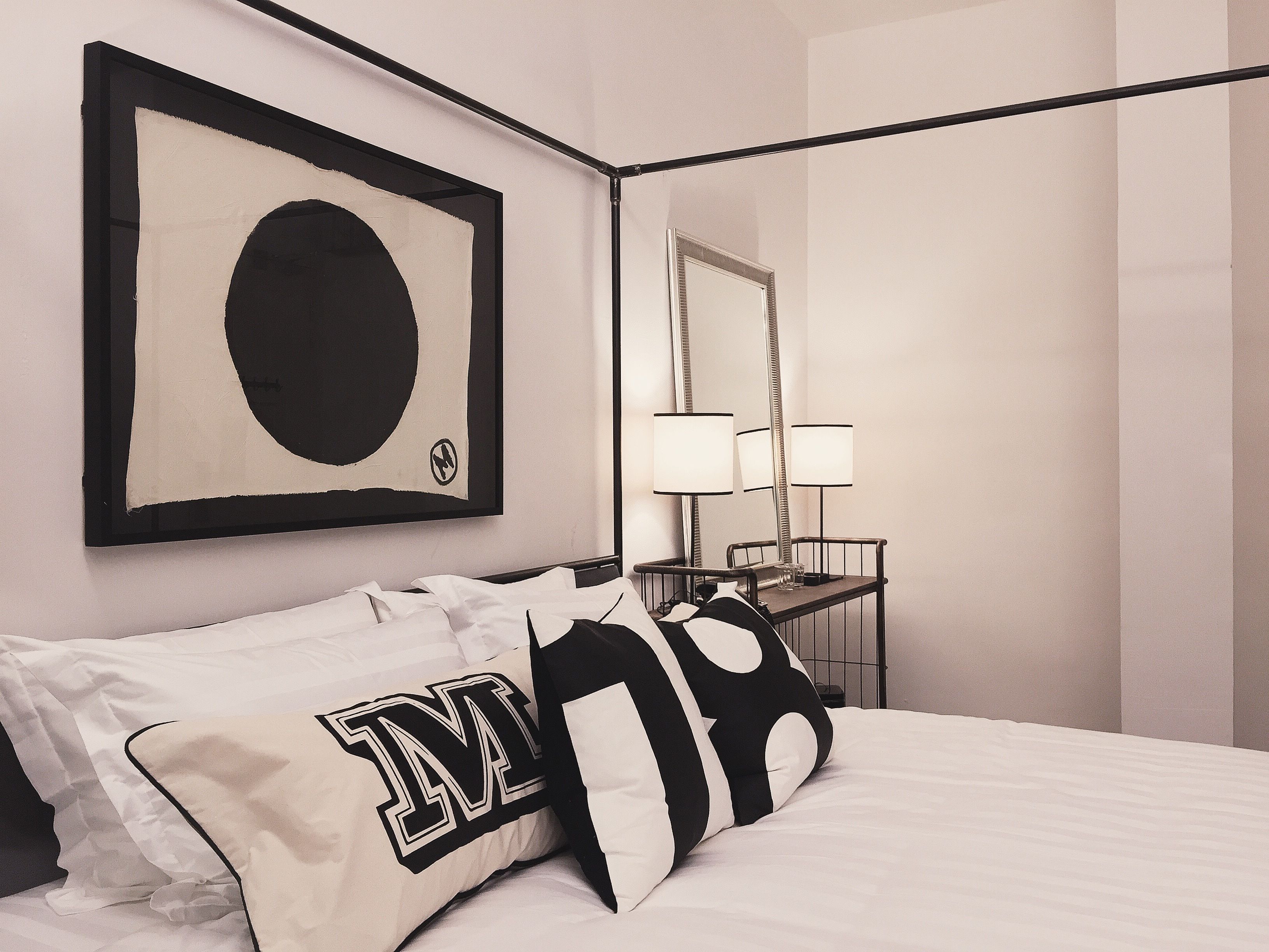 A neatly made bed with white and black bedding, a framed artwork above it, and a bedside table with a lamp.