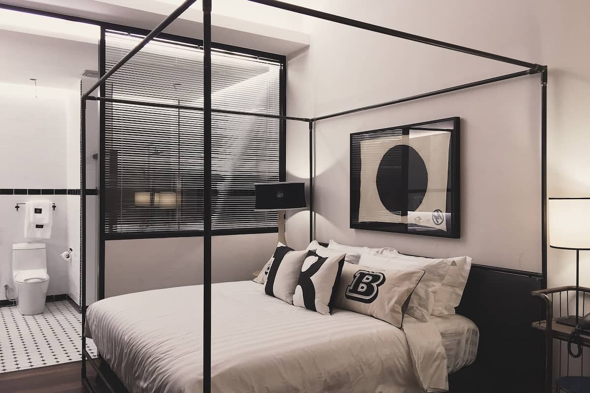 A modern bedroom with a four-poster bed, a large window with blinds, a bedside table with a lamp, and a wall-mounted mirror.