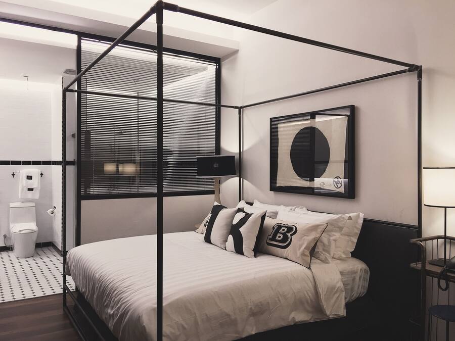 A modern bedroom with a four-poster bed, a large window with blinds, a bedside table with a lamp, and a wall-mounted mirror.
