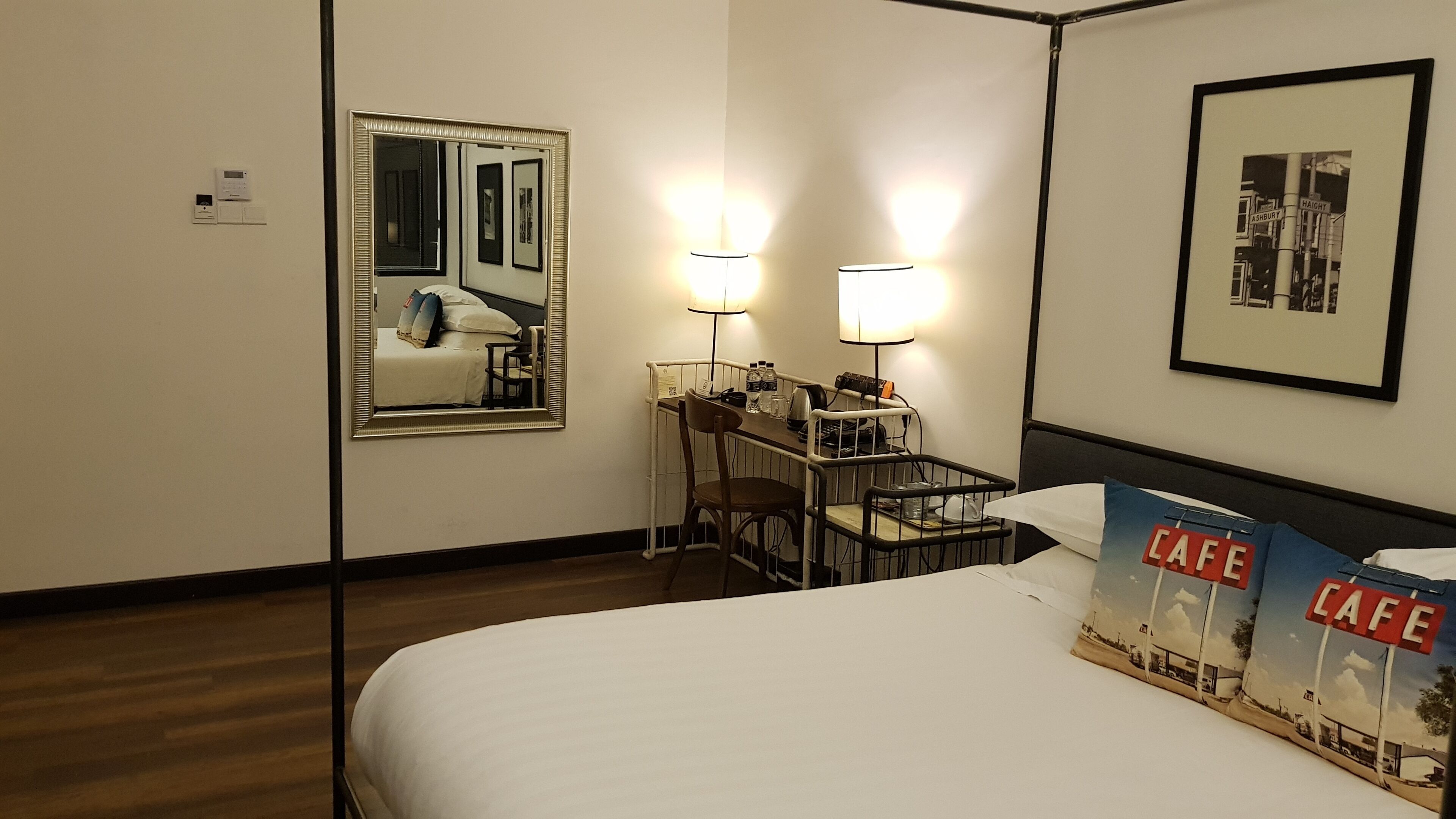 A hotel room with a bed, a desk with a chair, a mirror, two wall-mounted lamps, and a framed picture on the wall.