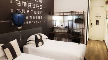 Habitación de hotel con dos camas, un reloj de pared grande, un perchero metálico con diversos objetos y una puerta con un cartel.