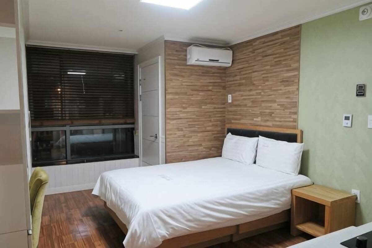 A bedroom with a bed, a window with blinds, an air conditioning unit, and a wooden nightstand.