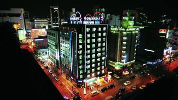 Ein beleuchtetes Stadtbild bei Nacht mit Häusern, einer belebten Straße und einem markanten Hotel mit Neonreklame.