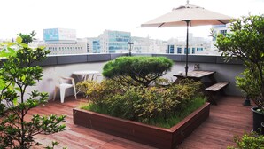 Una terraza en la azotea con piso de madera, una jardinera con plantas y una mesa con sillas bajo un amplio paraguas.