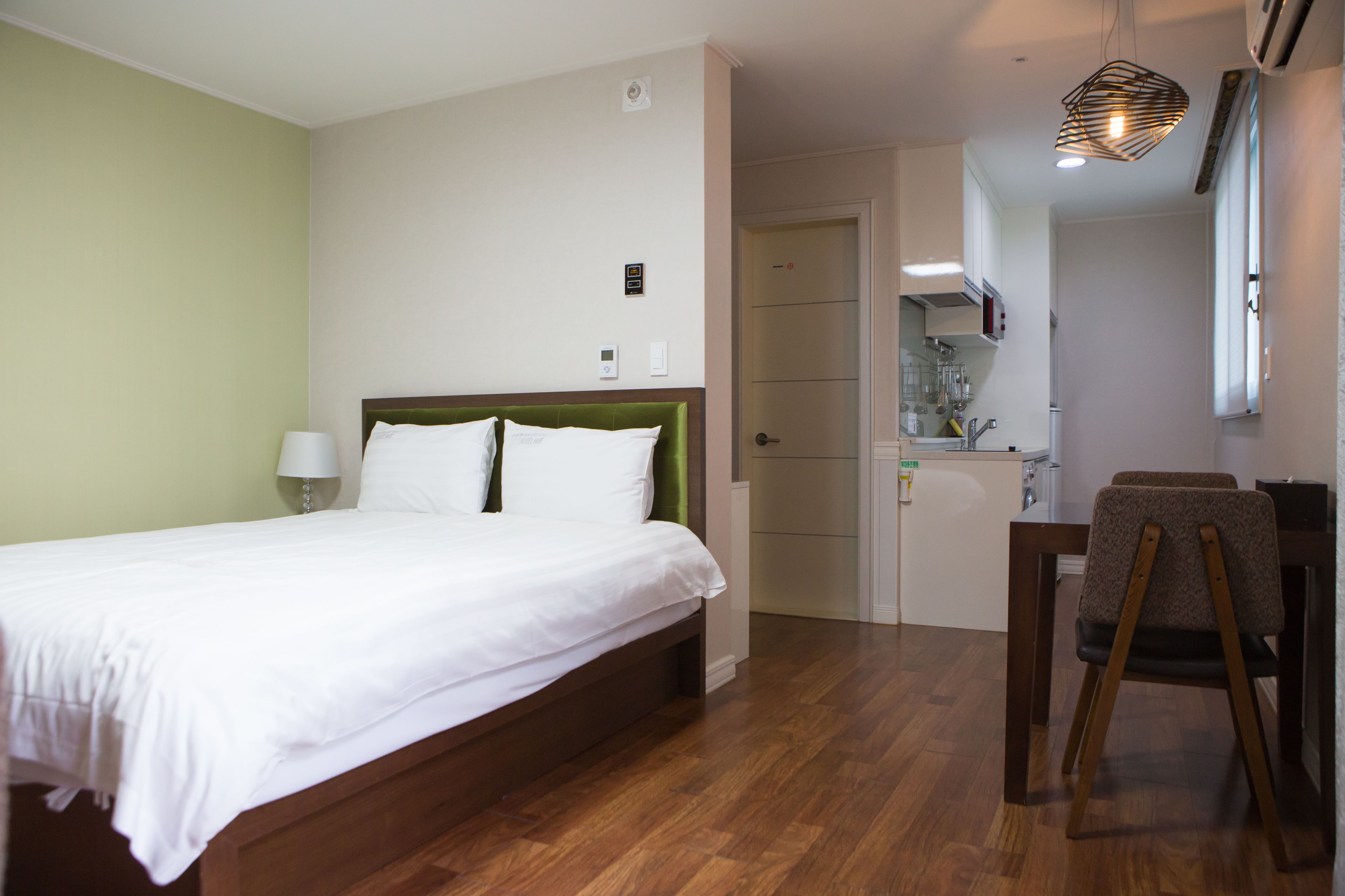 A hotel room with a bed, a desk, and a chair. There is a door leading to a kitchenette.