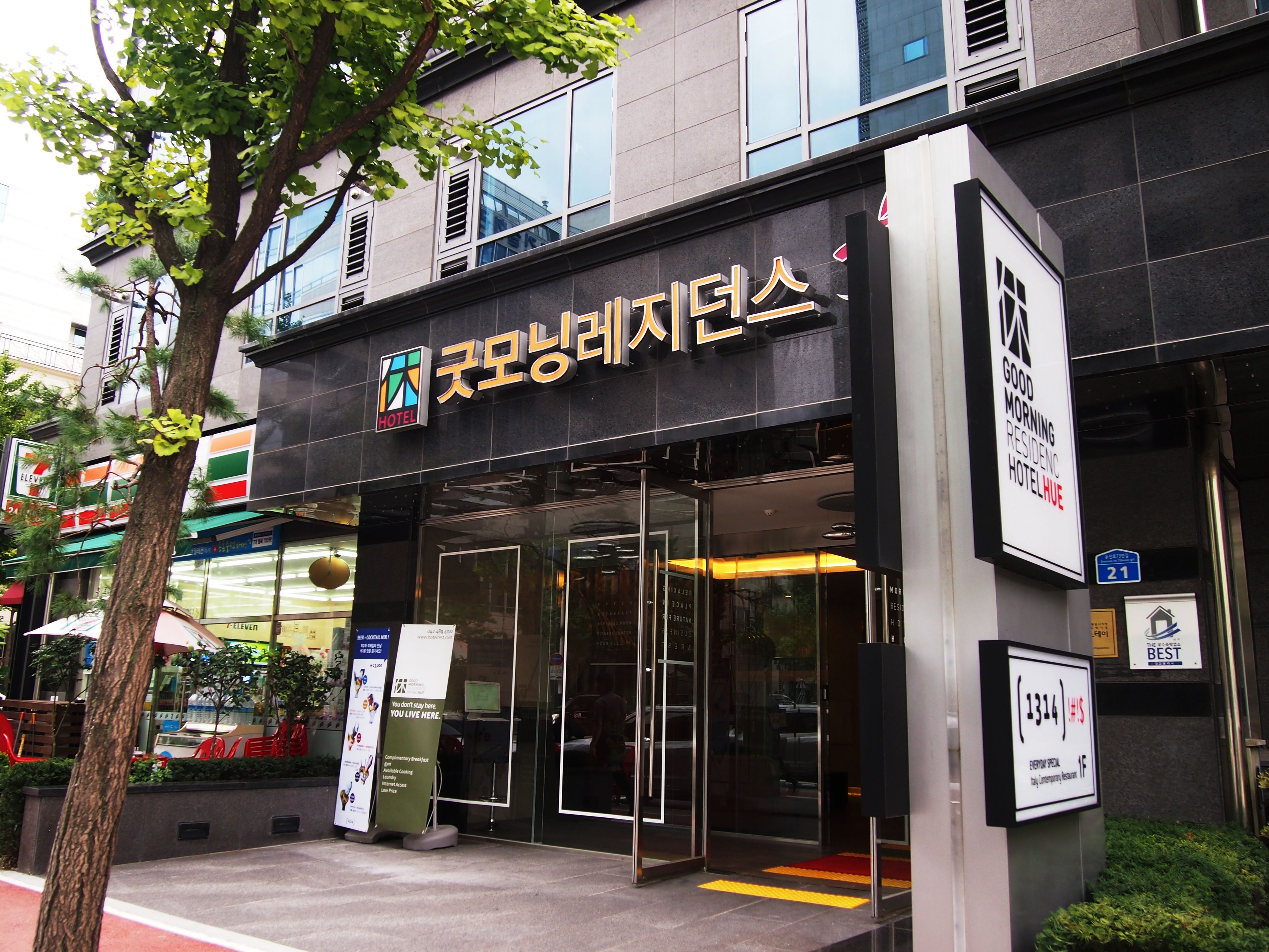 A hotel entrance with glass doors, a signboard with hotel name, and a 7-Eleven store nearby.