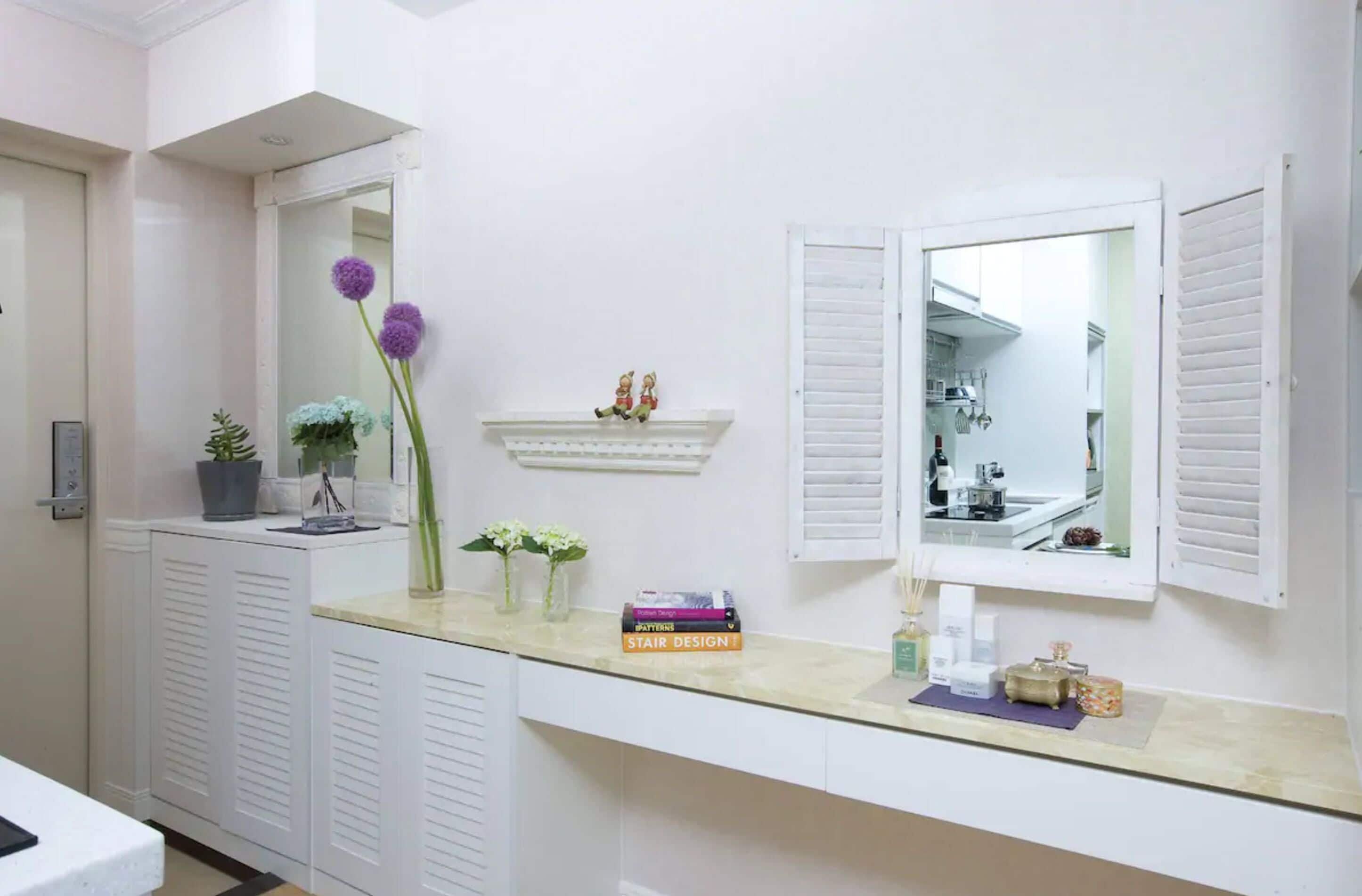 A bathroom with a vanity, a mirror, a shelf with decorative items, and a door leading to a kitchen.