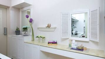 A bathroom with a vanity, a mirror, a shelf with decorative items, and a door leading to a kitchen.