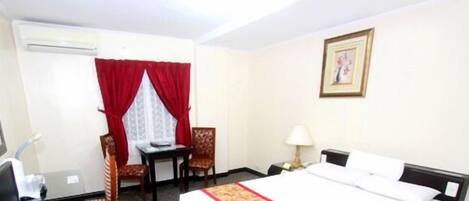 A hotel room with a bed, a desk with a chair, a lamp, and a window with red curtains.