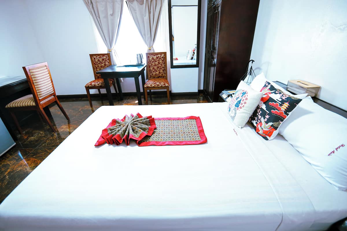 Cama bem arrumada com cabeceira decorativa em vermelho e bege, uma mesinha com cadeiras, e uma janela com cortinas.