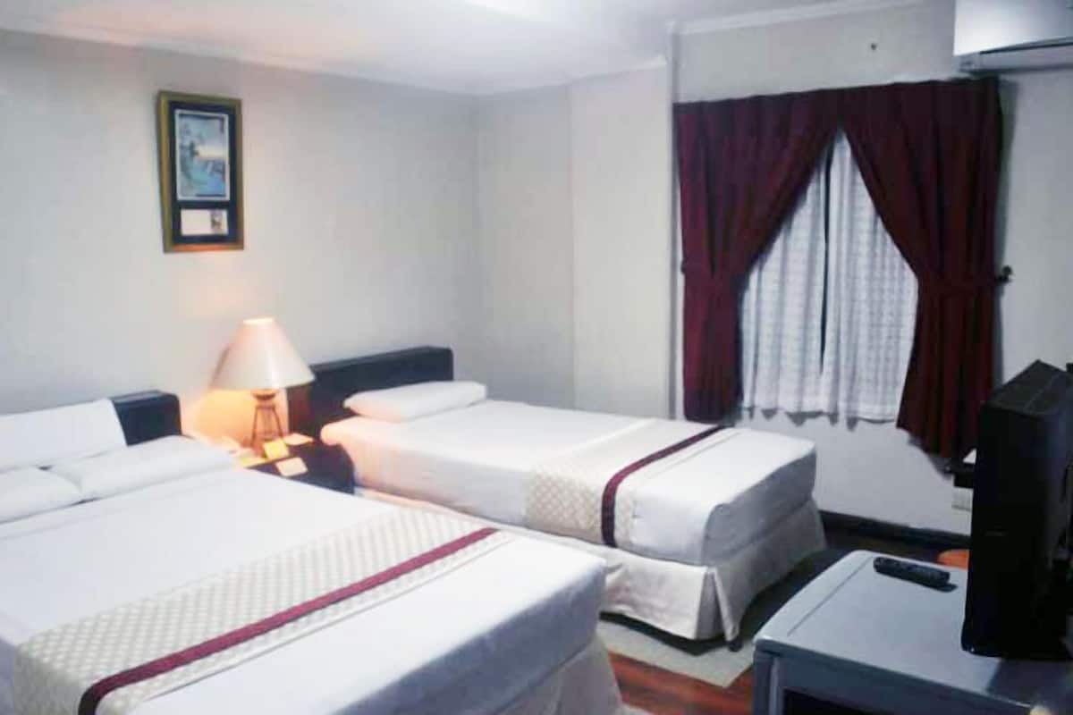 Quarto de hotel com duas camas de solteiro, uma mesa de trabalho com cadeira, uma luminária e uma janela com cortinas.