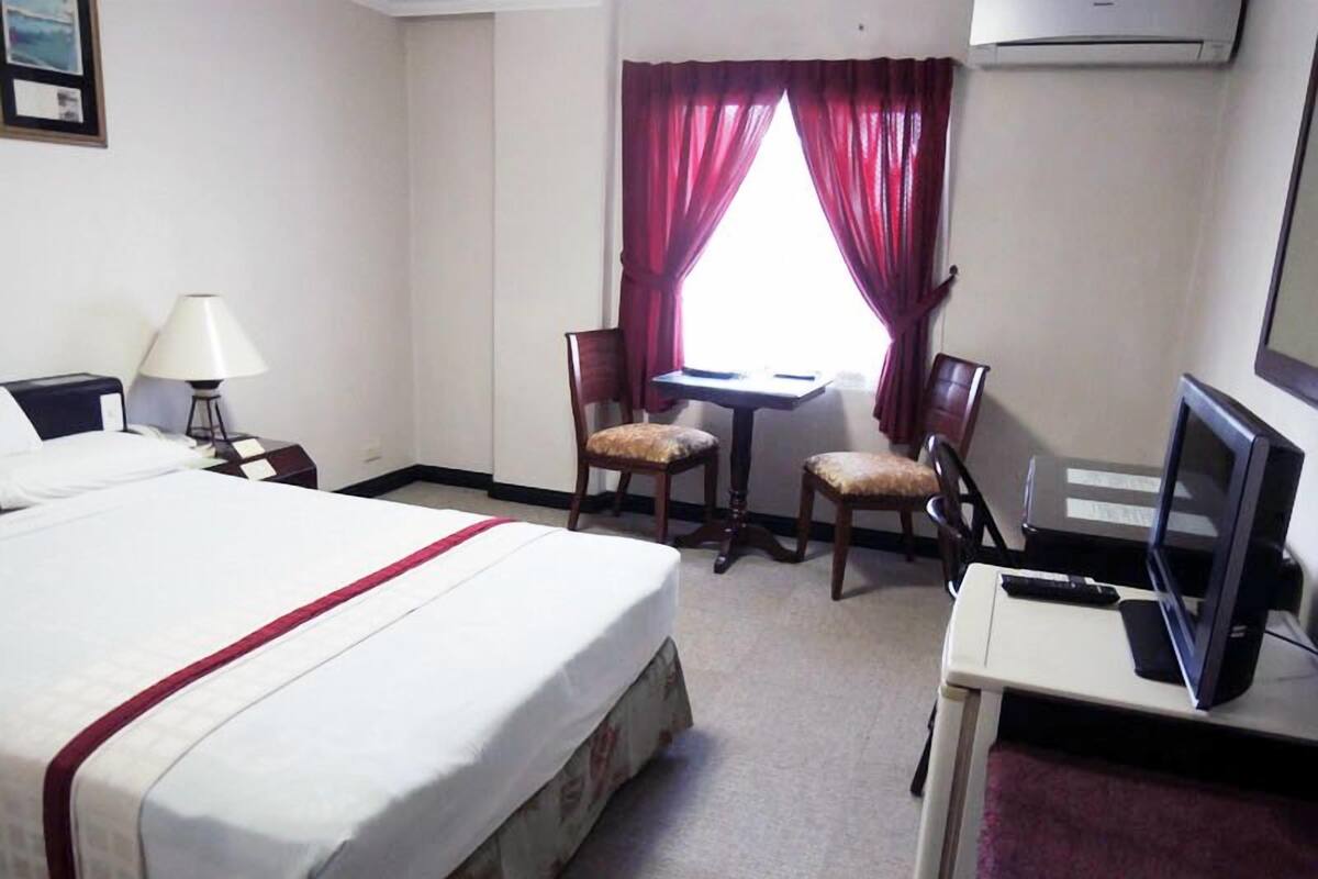 Quarto de hotel com cama, escrivaninha, duas cadeiras, televisão e luminária.