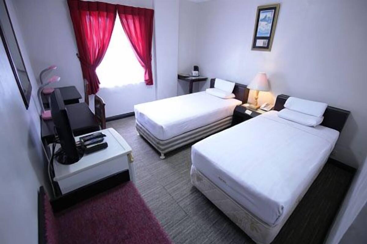 Quarto de hotel com duas camas, uma escrivaninha com cadeira, uma luminária de mesa, uma mesinha e uma janela com cortinas vermelhas.