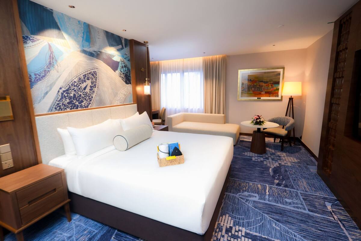 Een hotelkamer met een groot bed, een bureau, een stoel, een klein tafeltje met een lamp, en een schilderij aan de muur.