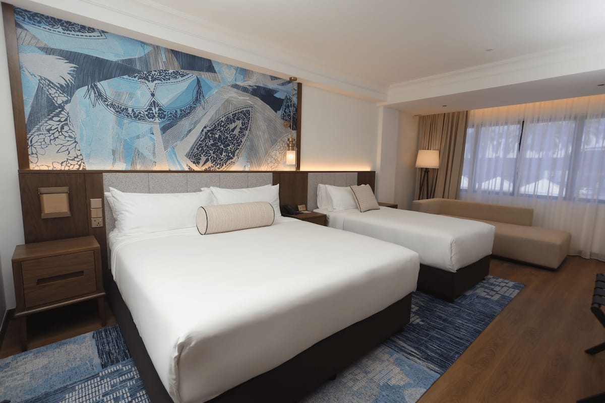 Een hotelkamer met twee bedden, een groot abstract schilderij, een nachtkastje, een lamp en uitzicht op het buiten.