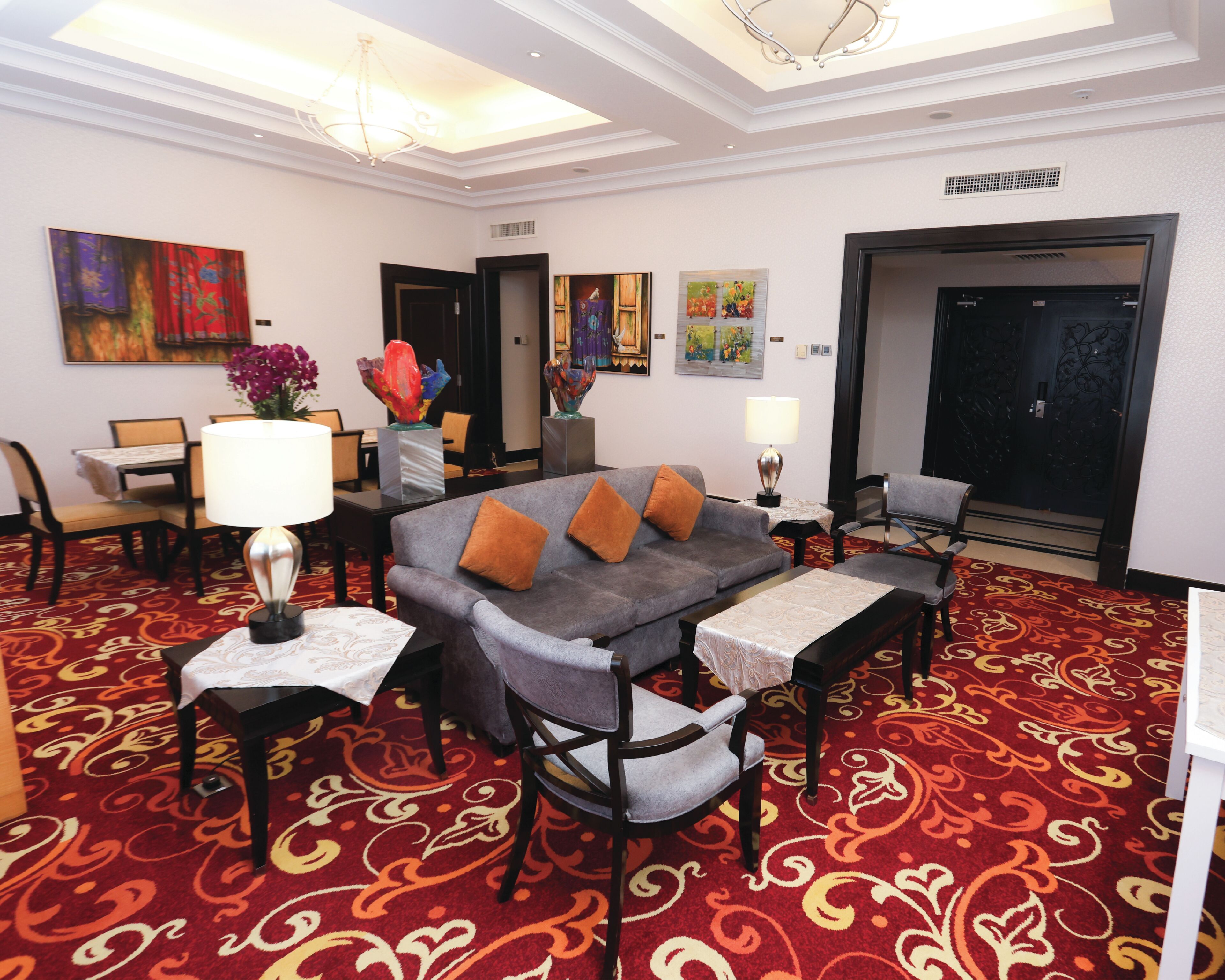A hotel room with a grey sofa, two armchairs, a dining table with chairs, a chandelier, and a red patterned carpet.