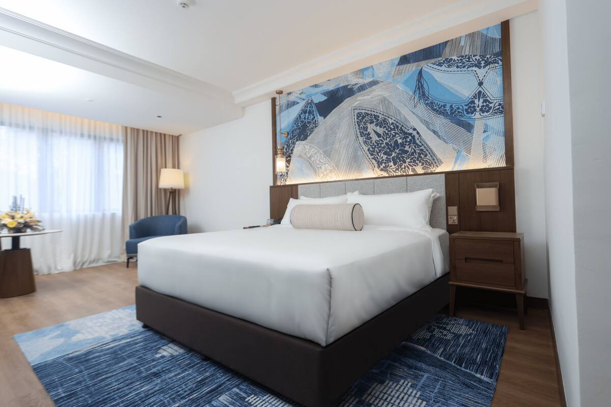 Hotelkamer met een groot bed, nachtkastje, bureau met lamp, stoel en een groot schilderij aan de muur.