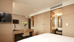A modern hotel room with a large bed, a desk with a chair, a flat-screen TV, a vase with flowers, and a large mirror. - Mayplace Seoul (Seoul)