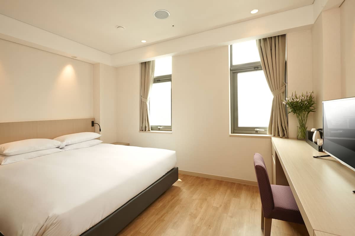 A hotel room with a bed, a desk, a chair, a TV, and two windows with curtains.