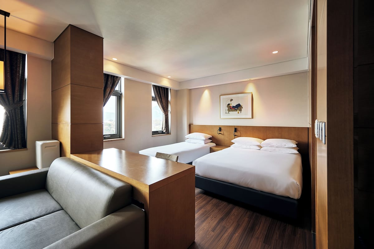 A hotel room with a sofa, two beds, a wooden headboard, a framed picture, and a window with curtains.