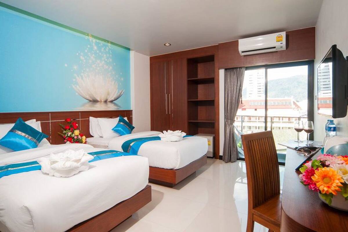 A hotel room with two beds, a desk, a TV, and a large window with a view of buildings.