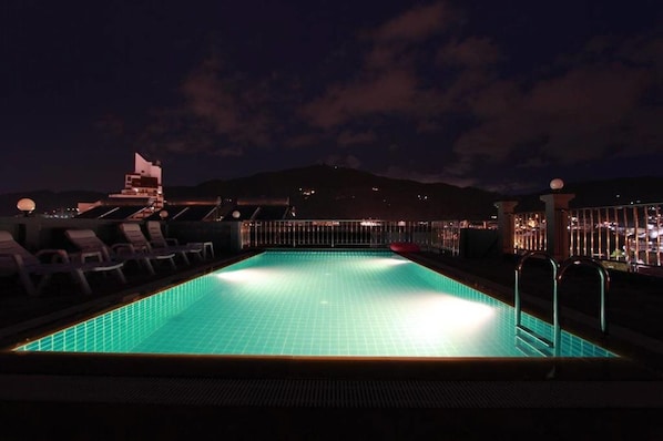 Una piscina rectangular con piso de azulejos, rodeada de sillas de descanso y una barandilla.