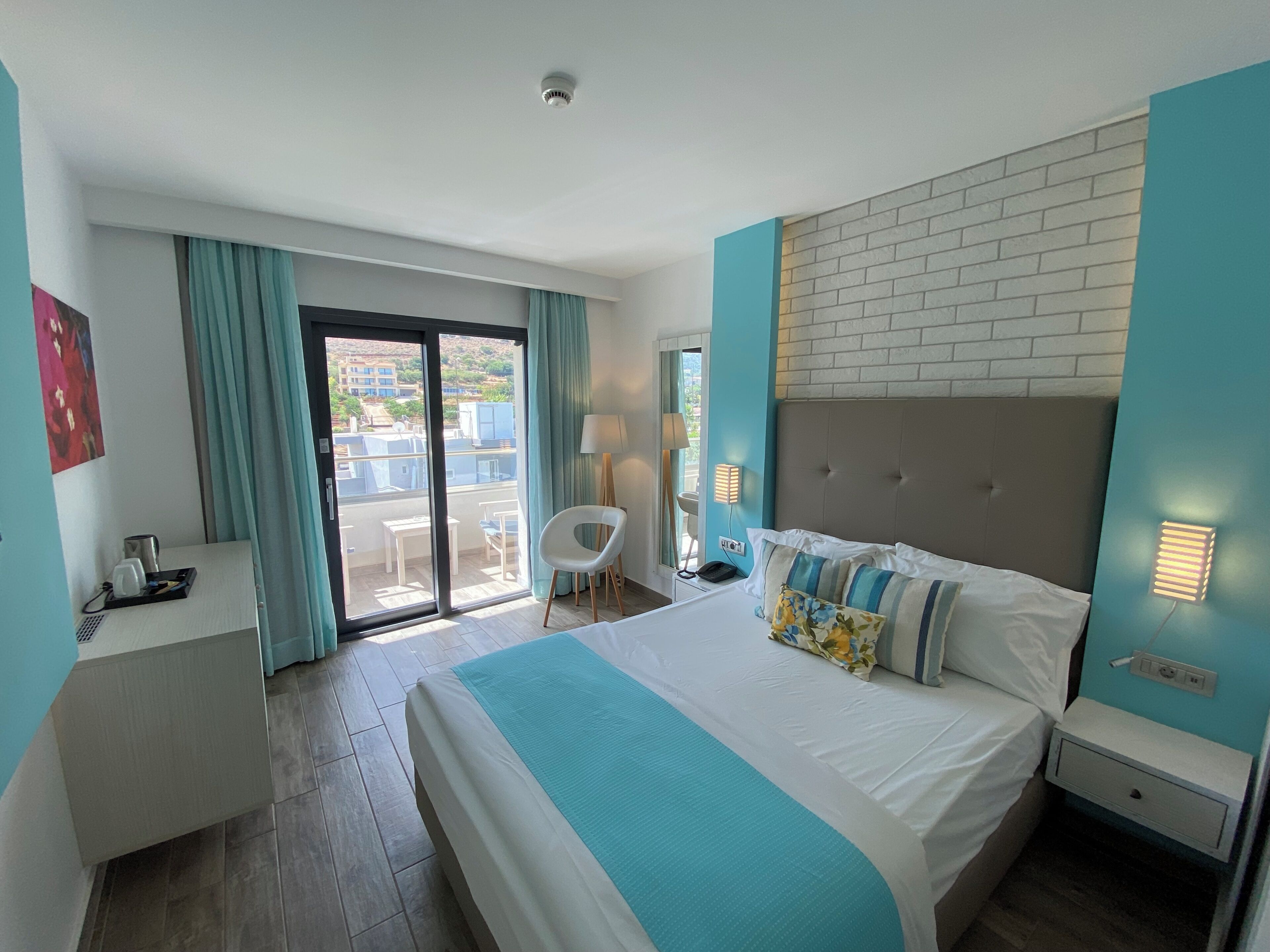 Habitación de hotel con cama, escritorio, silla, balcón con mesa y sillas, y vista a edificios.