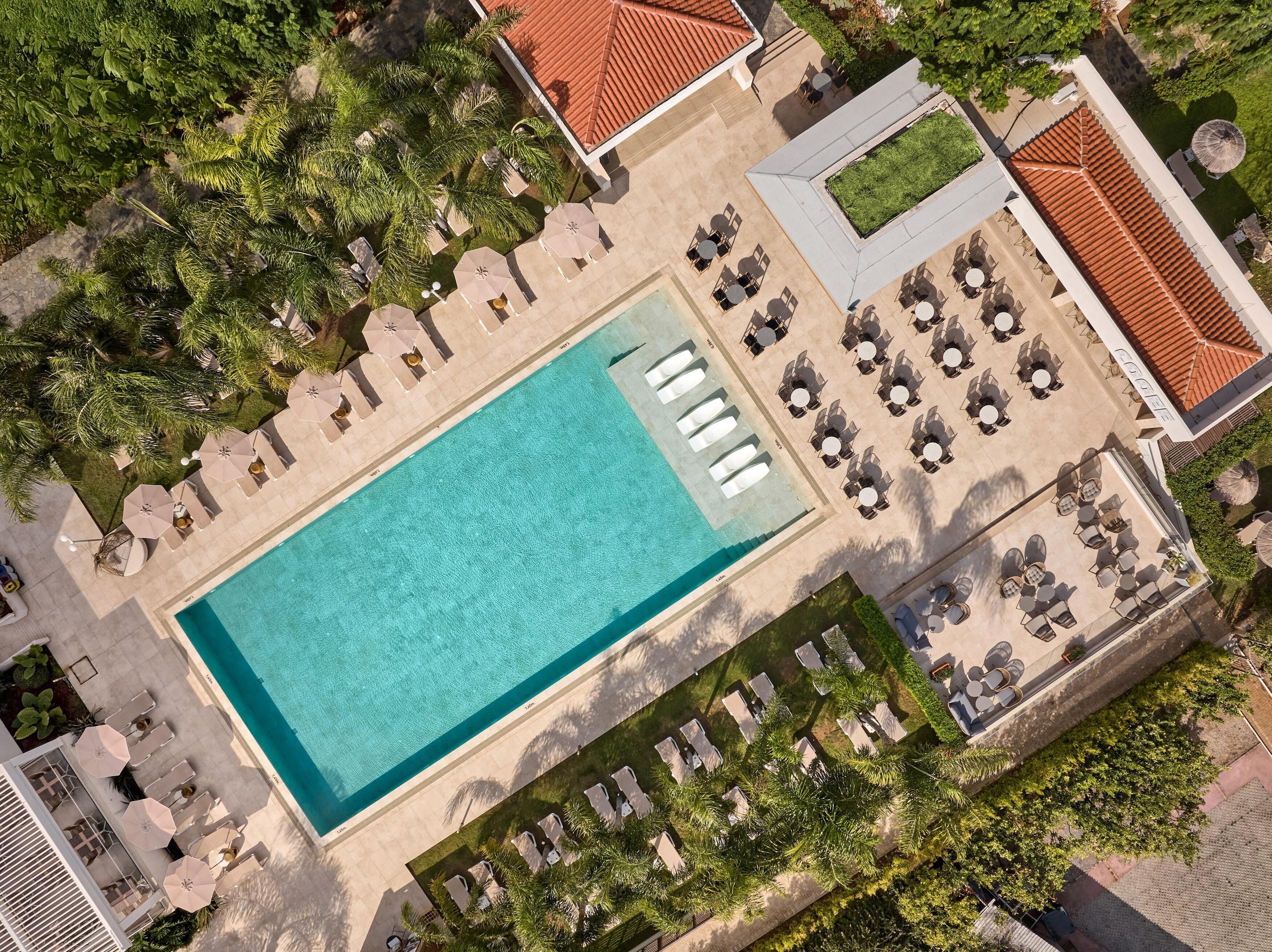 An aerial view of a resort with a large swimming pool surrounded by sun loungers and umbrellas, a covered outdoor seating area, and lush greenery.