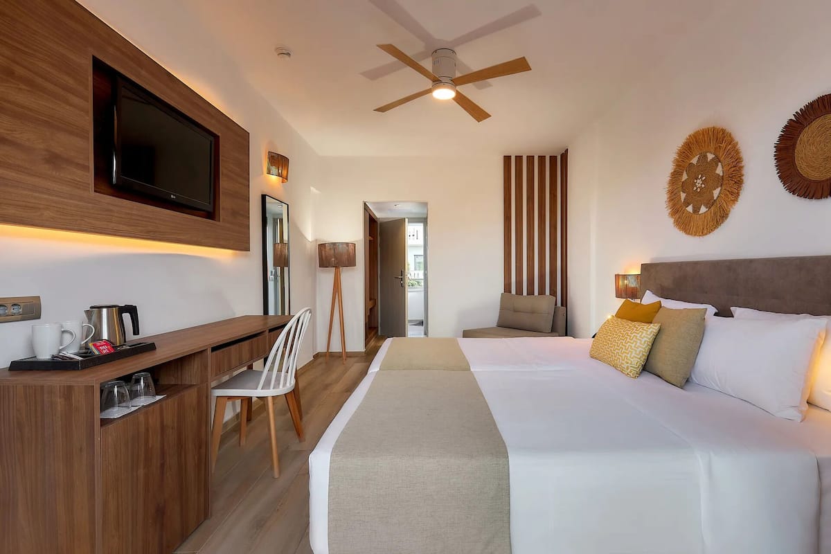 A hotel room with a bed, bedside table, TV, ceiling fan, and a view of the outside.