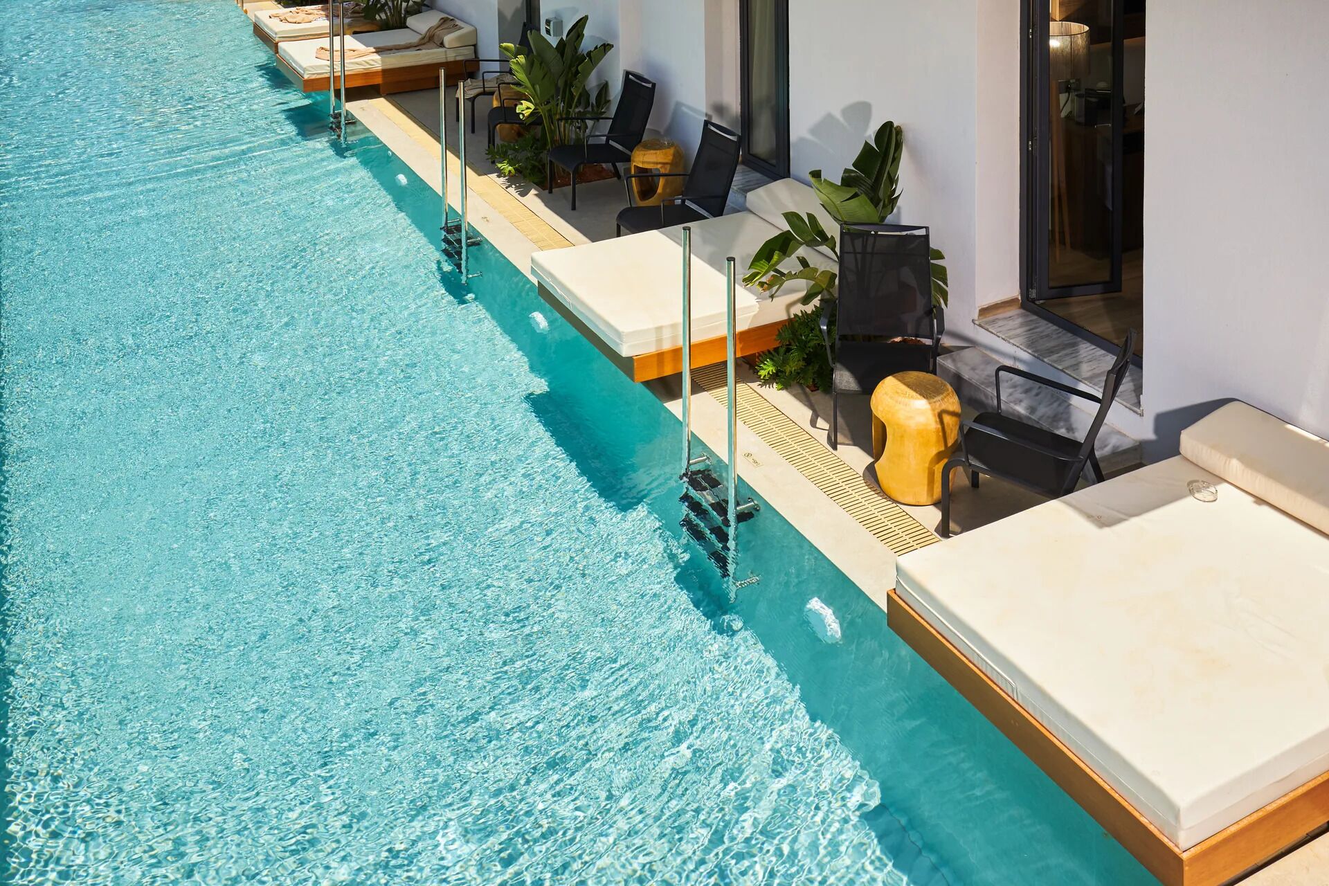 Una piscina larga con sillas de descanso y mesas a lo largo del borde, un muelle de madera y un edificio moderno con grandes ventanales.