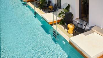 Una piscina larga con sillas de descanso y mesas a lo largo del borde, un muelle de madera y un edificio moderno con grandes ventanales.