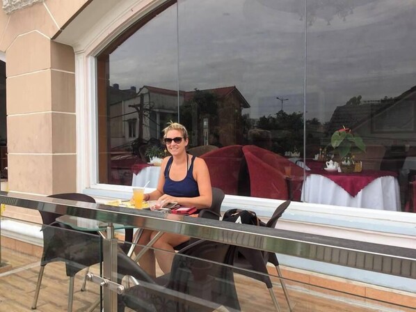 Una persona sentada en una mesa al aire libre, con un vaso de bebida, una silla roja y una mesa con mantel blanco.