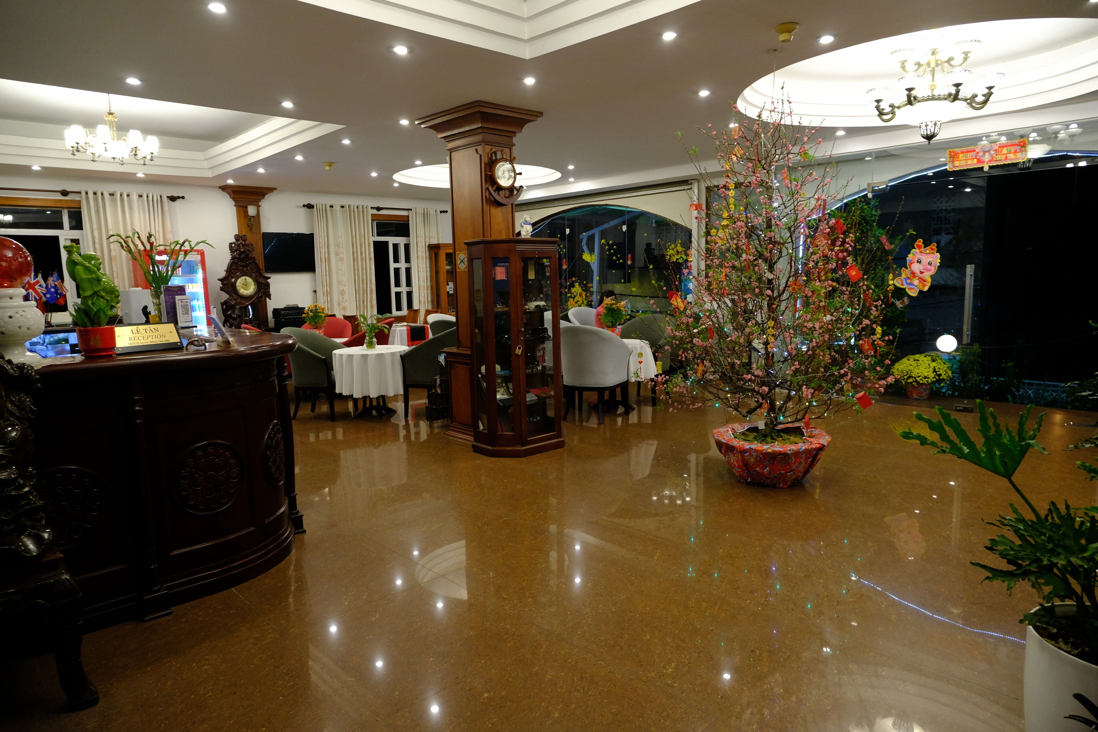 A hotel lobby with a reception desk, a clock, a chandelier, and potted plants.