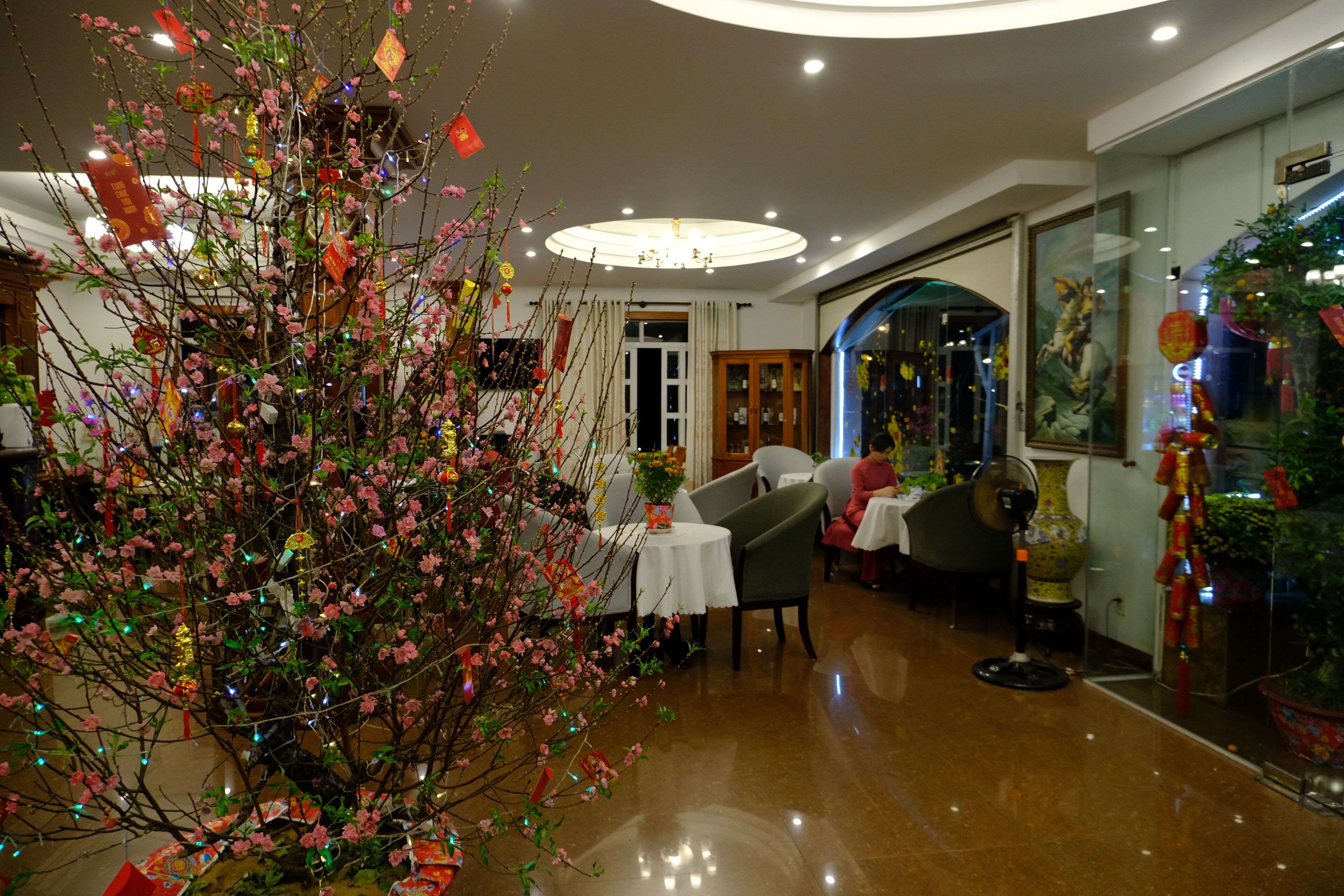 Un'ampia hall d'albergo con un grande albero artificiale, tavoli, sedie e un vaso decorativo tradizionale.
