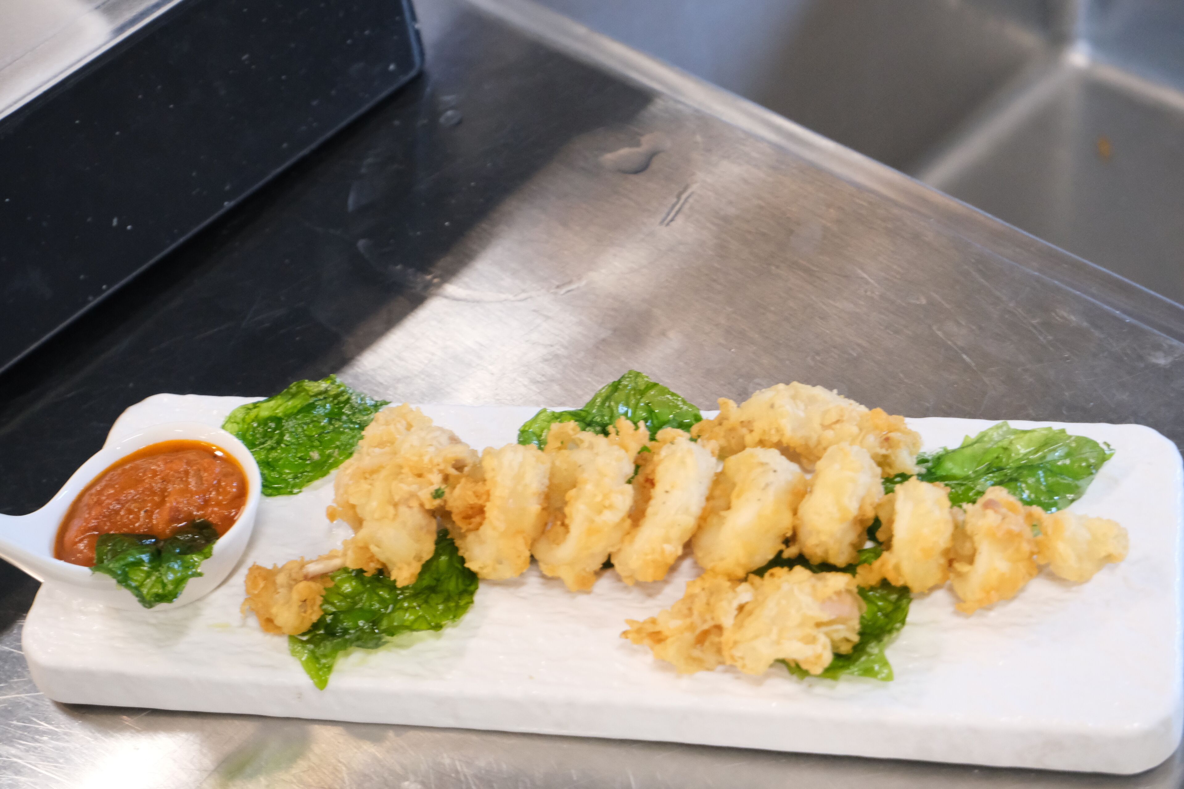 A white rectangular plate with fried food and a small bowl of sauce, placed on a stainless steel surface.