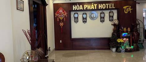 Un hall d'accueil d'hôtel avec une enseigne indiquant « HÔTEL HOA PHÁT », un comptoir d'accueil en bois, une horloge et une décoration murale traditionnelle.