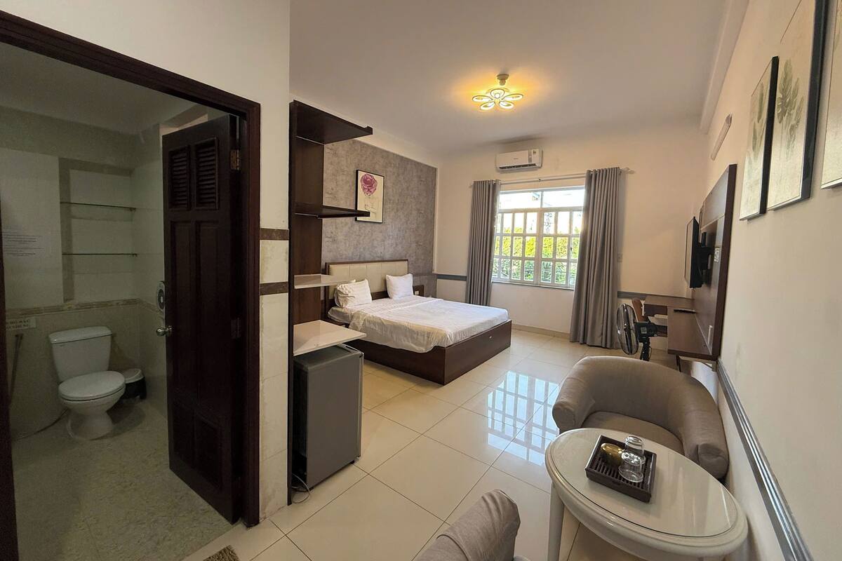 A hotel room with a bed, a small table, a chair, a ceiling fan, a window with curtains, and a door leading to a bathroom.