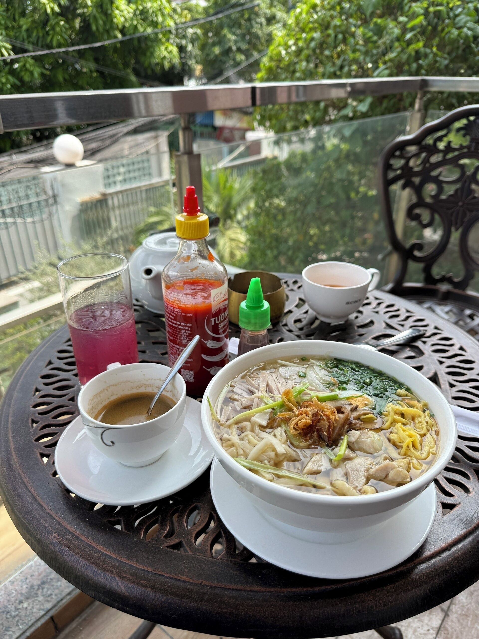 Una mesa con un tazón de sopa de fideos, una taza de café, un vaso de bebida helada y salsas.
