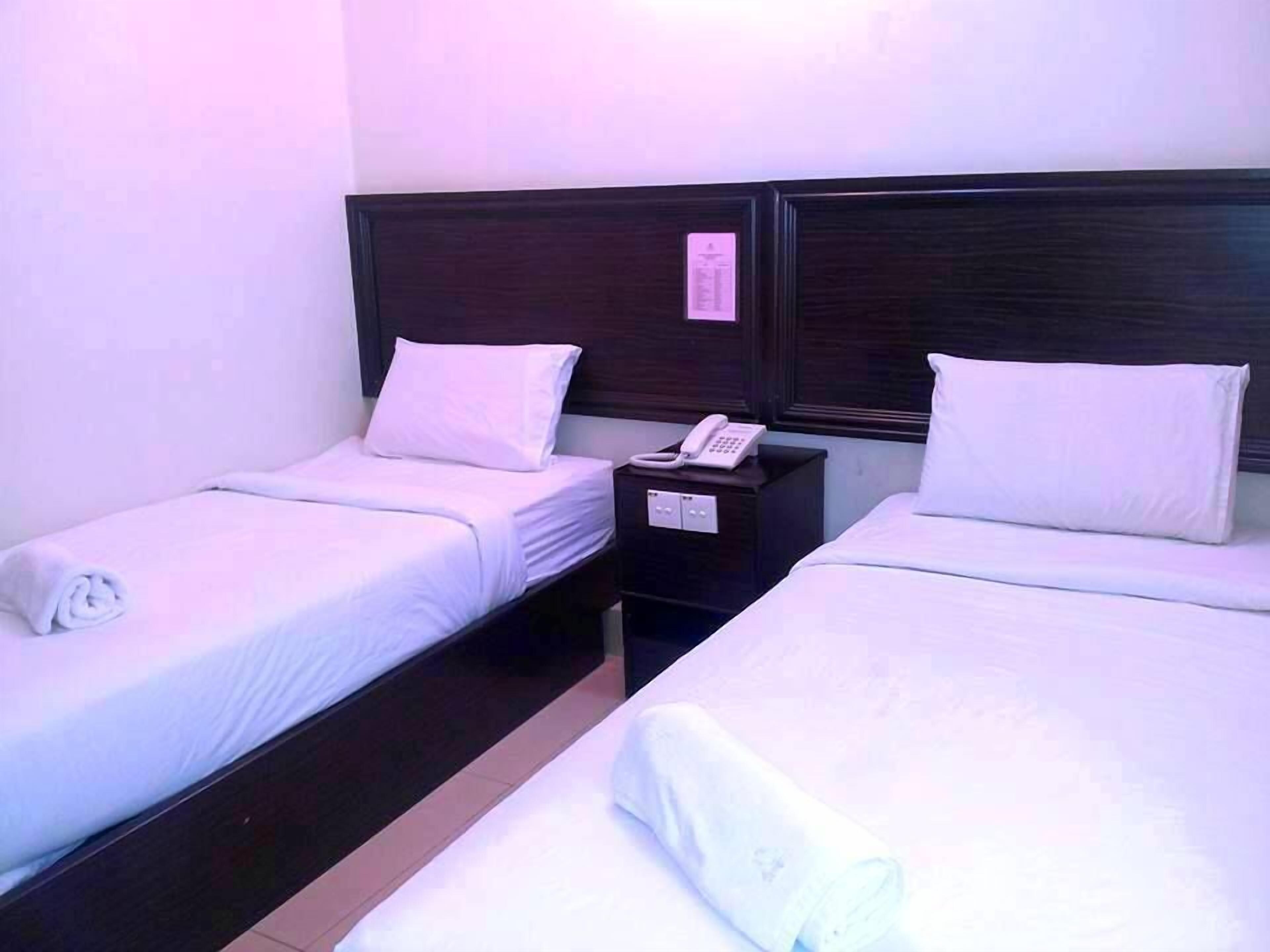 Habitación de hotel con dos camas individuales, cada una con sábanas y almohadas blancas, un cabecero de madera oscura y una mesita de noche con un teléfono y una tarjeta pequeña.