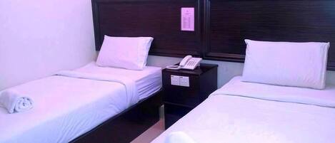 Habitación de hotel con dos camas individuales, cada una con sábanas y almohadas blancas, un cabecero de madera oscura y una mesita de noche con un teléfono y una tarjeta pequeña.