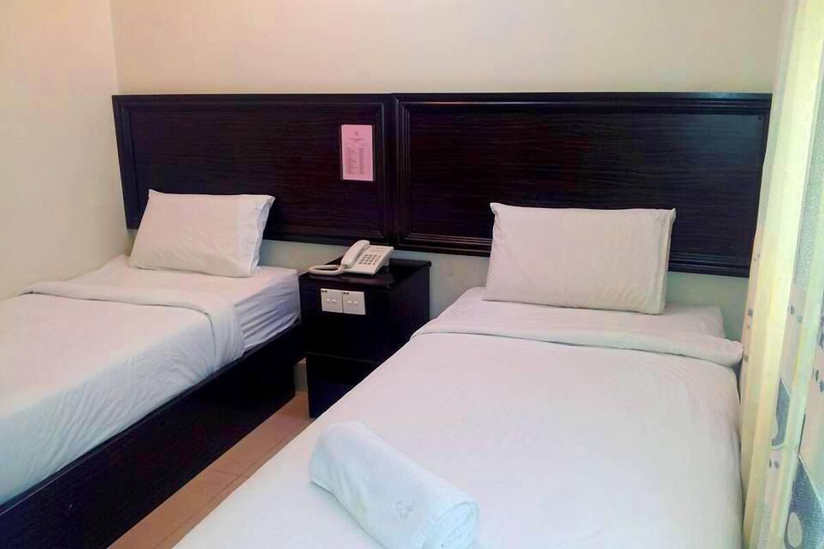 Ein Hotelzimmer mit zwei Einzelbetten, jeweils mit Kissen und einer weißen Decke. Es gibt einen Nachttisch mit Telefon und einem kleinen Schrank.