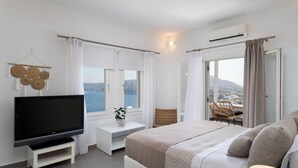 A modern hotel room with a large bed, a flat-screen TV on a white stand, a small side table, a wicker chair, and a view of the sea through the window. - The Zen Villa (Santorini)