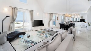 A spacious living room with a large sofa, a glass coffee table, and a flat-screen TV. There is a dining area with a kitchen in the background. - The Zen Villa (Santorini)