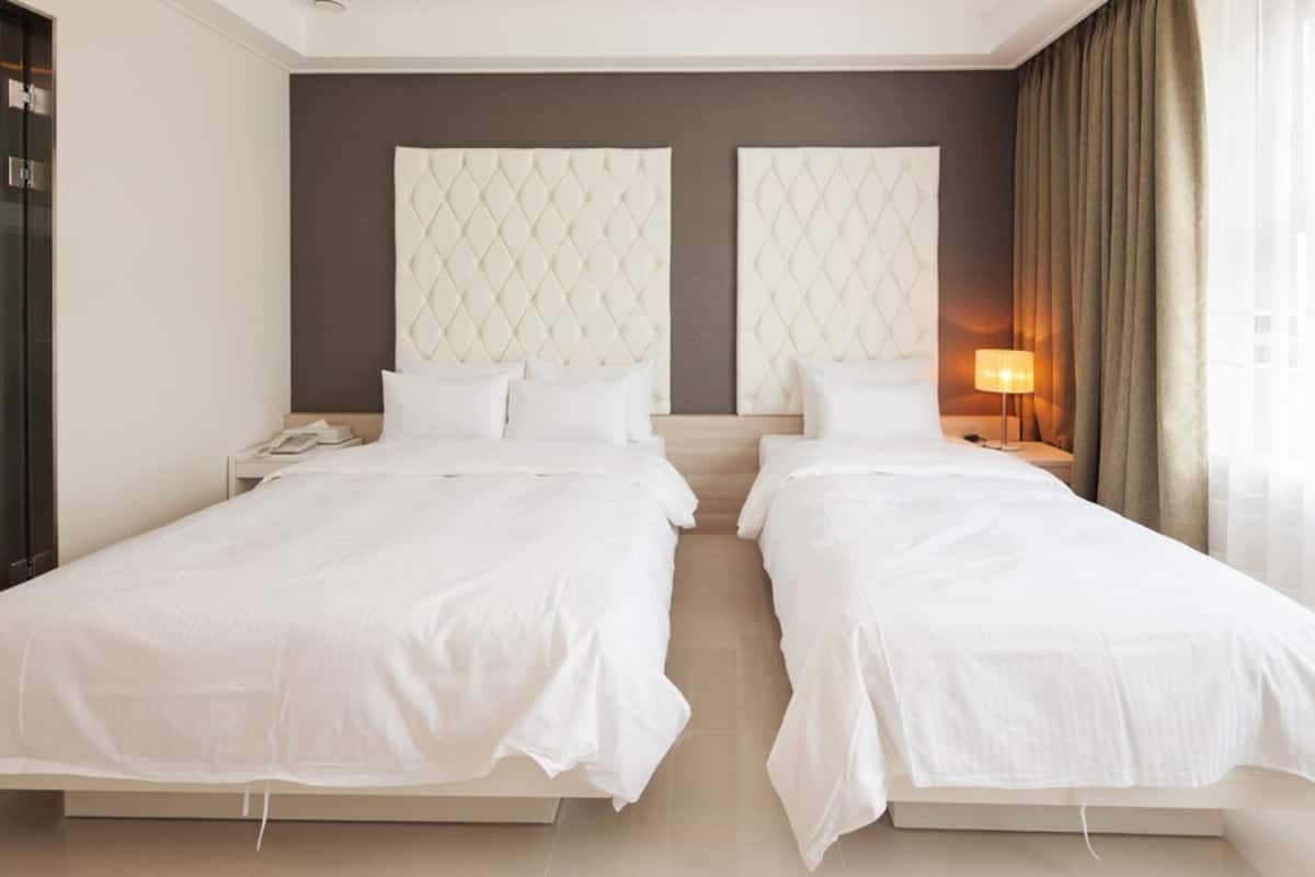 A hotel room with two beds, a large window with curtains, and a nightstand with a lamp.