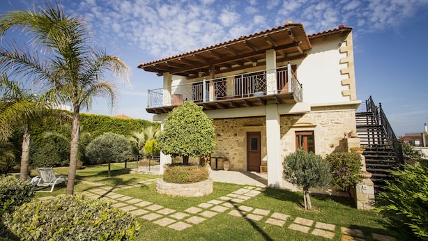 A two-story house with a stone facade, a balcony, and a well-maintained garden with palm trees and a circular hedge. - Secreto Studios (Kissamos)
