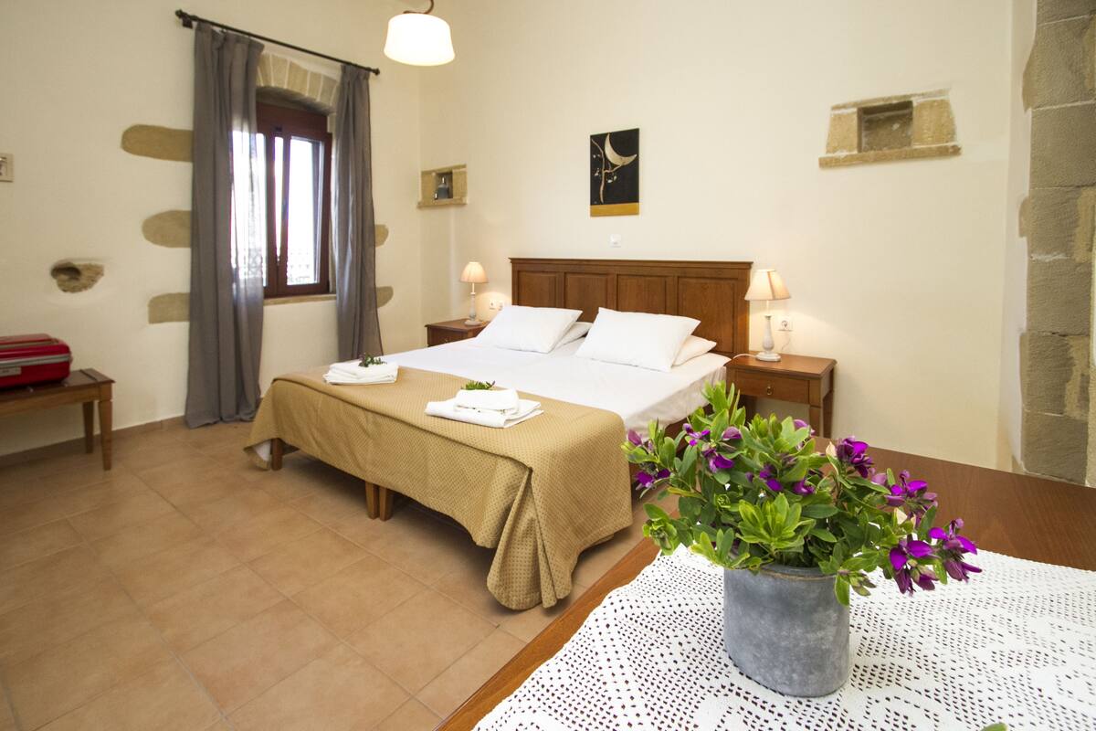 A hotel room with a bed, bedside tables, a window with curtains, a wall-mounted artwork, and a potted plant on a table.