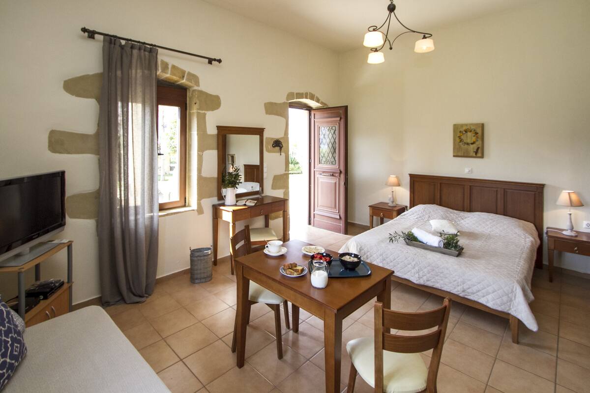 A hotel room with a bed, a TV, a dining table with breakfast items, a mirror, and a window with curtains.