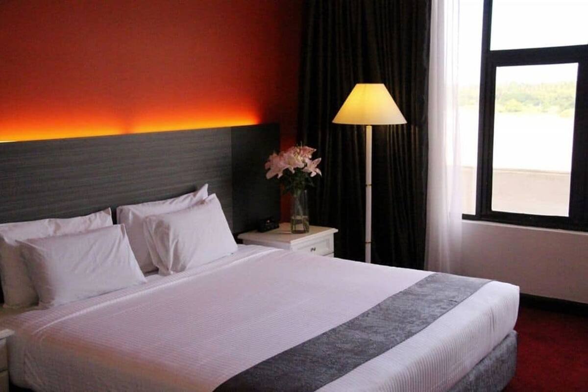 Ein Hotelzimmer mit Bett, weißen Kissen, einem Nachttisch, einer Lampe und einem Fenster mit Blick.