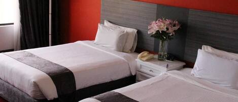 A hotel room with two beds, a bedside table with a lamp, and a vase with flowers.
