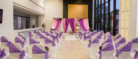 A ceremony aisle with rows of chairs, each with a white cover and purple sashes, leading to a stage with pink drapes.